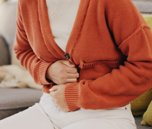 Stomach ache, cramps and hands of woman with abdomen pain due to constipation, menstruation or ibs issue. Sick, home and person suffering and holding belly in a house lounge, couch and living room.